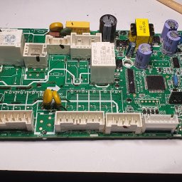 Alex Crisard - Płyta główna urządzenia elektronicznego z widocznymi elementami, takimi jak kondensatory i złącza, leżąca na białym tle. Obok pędzel malarski.