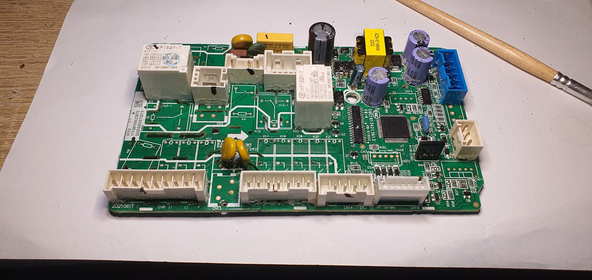 Płyta główna urządzenia elektronicznego z widocznymi elementami, takimi jak kondensatory i złącza, leżąca na białym tle. Obok pędzel malarski.