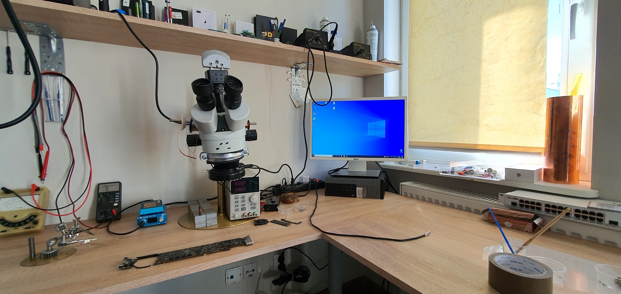 Stanowisko pracy serwisanta elektroniki z mikroskopem, multimetrem i płytą główną. Widoczne narzędzia i monitor. Jasne oświetlenie, schludne otoczenie.