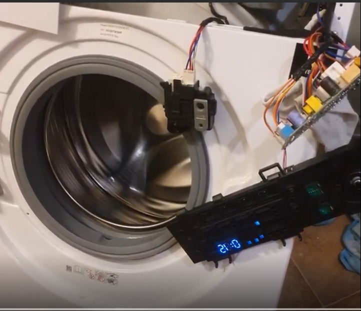 Otwarta pralka z widocznym bębnem i odłączonym panelem sterowania z podświetlonym wyświetlaczem, sugerująca naprawę lub serwis urządzenia.