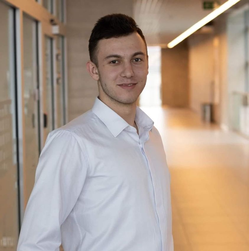 Młody mężczyzna w białej koszuli, uśmiechnięty, w nowoczesnym biurze z przeszklonymi ścianami i jasnym oświetleniem. Portret biznesowy.