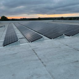 Smart team - Rząd paneli fotowoltaicznych na płaskim dachu w pochmurny dzień. Widoczne mocowania paneli i odległy krajobraz o zachodzie słońca w tle.
