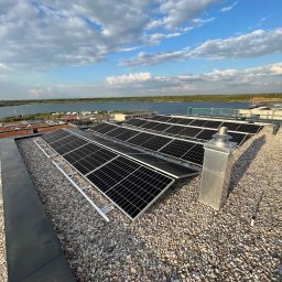 Smart team - Instalacja fotowoltaiczna na dachu pokrytym żwirem, z widokiem na jezioro i błękitne niebo. Panele ustawione rzędami, widoczny komin wentylacyjny.