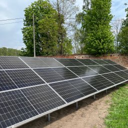 Smart team - Instalacja fotowoltaiczna na gruncie, panele ułożone rzędami, tło z zieleni i fragment ceglanego muru. Słoneczny dzień, widok z niskiej perspektywy.