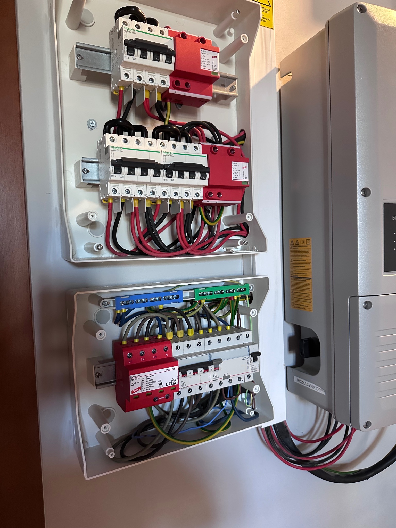 Rozdzielnia elektryczna z zabezpieczeniami nadprądowymi i przeciwprzepięciowymi, widoczne przewody w różnych kolorach oraz szyna DIN. Obok szara obudowa rozłącznika DC.