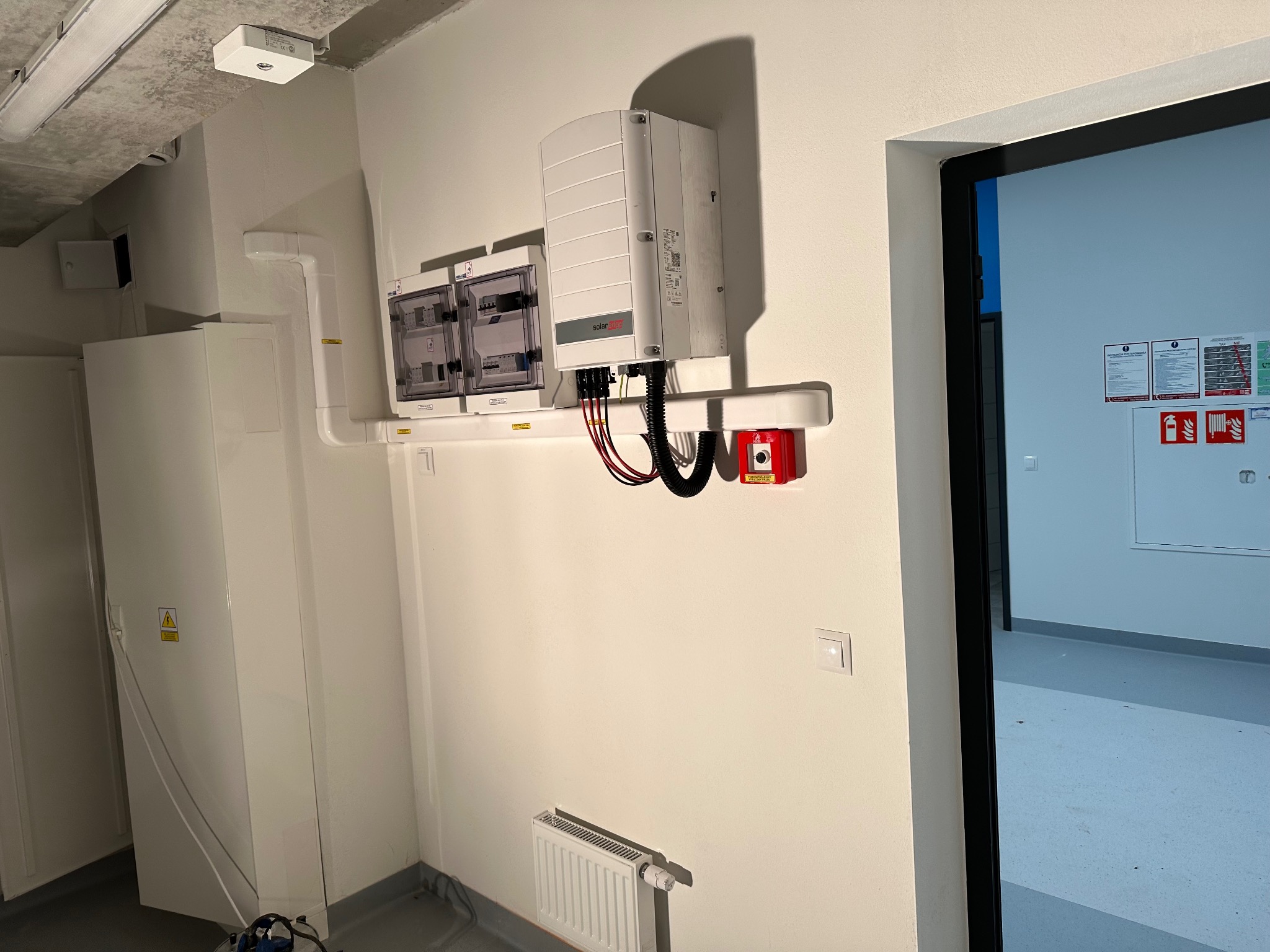 Białe skrzynki elektryczne i falownik SolarEdge zamontowane na ścianie wewnątrz budynku. Widoczny grzejnik i fragment pomieszczenia przez otwarte drzwi.