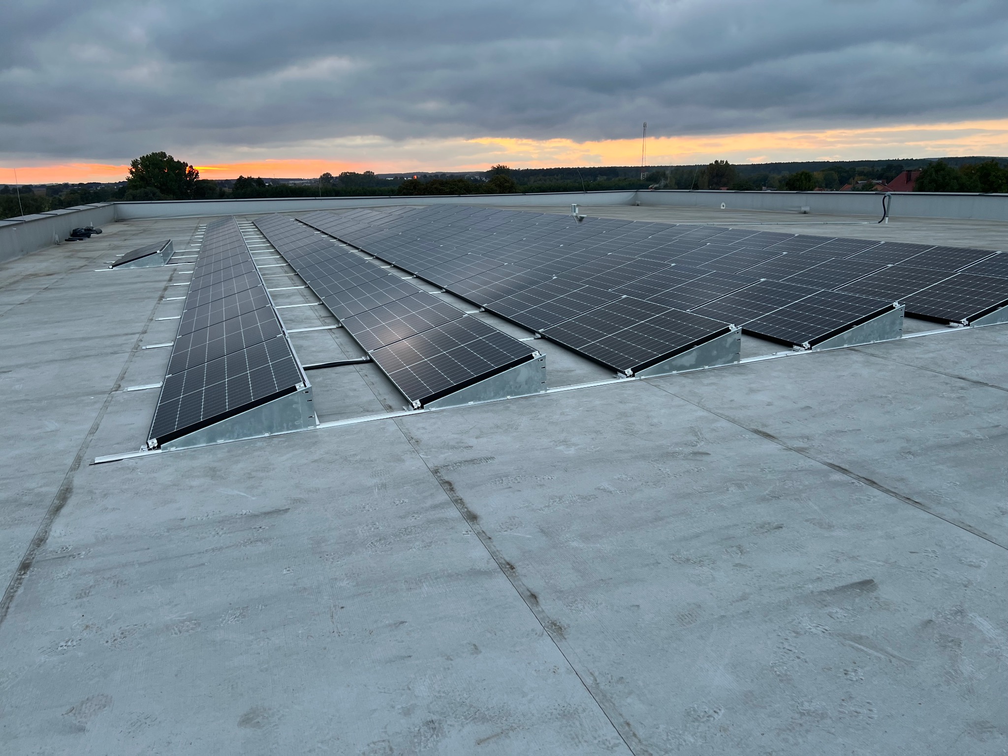 Rząd paneli fotowoltaicznych na płaskim dachu w pochmurny dzień. Widoczne mocowania paneli i odległy krajobraz o zachodzie słońca w tle.