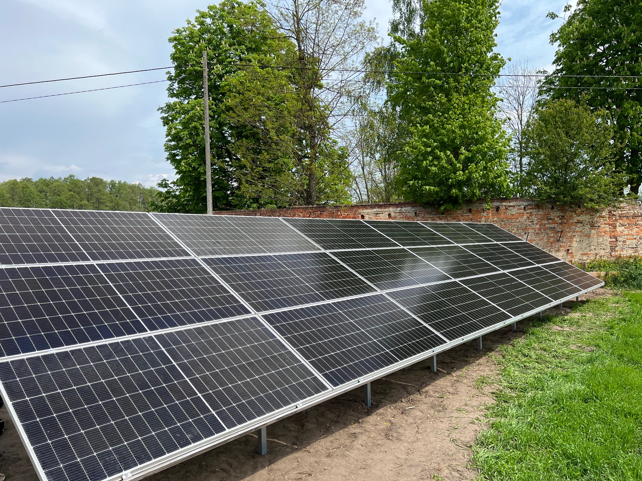 Instalacja fotowoltaiczna na gruncie, panele ułożone rzędami, tło z zieleni i fragment ceglanego muru. Słoneczny dzień, widok z niskiej perspektywy.