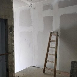 eMBUD - Pokój w trakcie szpachlowania ścian i sufitu, widoczna drewniana drabina oparta o ścianę. Surowy beton na podłodze i wystające kable z sufitu.