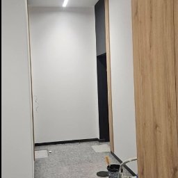 eMBUD - Korytarz w trakcie malowania: białe ściany, szara podłoga, drabina malarska, puszki z farbą. Widoczny znak ewakuacyjny i lampa sufitowa.