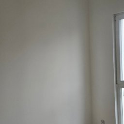 eMBUD - Jasne wnętrze po malowaniu: gładkie, kremowe ściany, drewnopodobna podłoga, biała listwa przypodłogowa i okno. Widoczne gniazdka elektryczne.