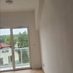 Jasne, odświeżone wnętrze z parkietem i dużym oknem balkonowym. Ściany pomalowane na biało, widoczny fragment sufitu i listwy przypodłogowe. Minimalistyczny styl.