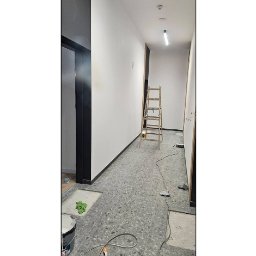 eMBUD - Wnętrze korytarza w trakcie odświeżania: pomalowane ściany na biało, szara podłoga z terazzo, drabina malarska, widoczne ślady prac remontowych.