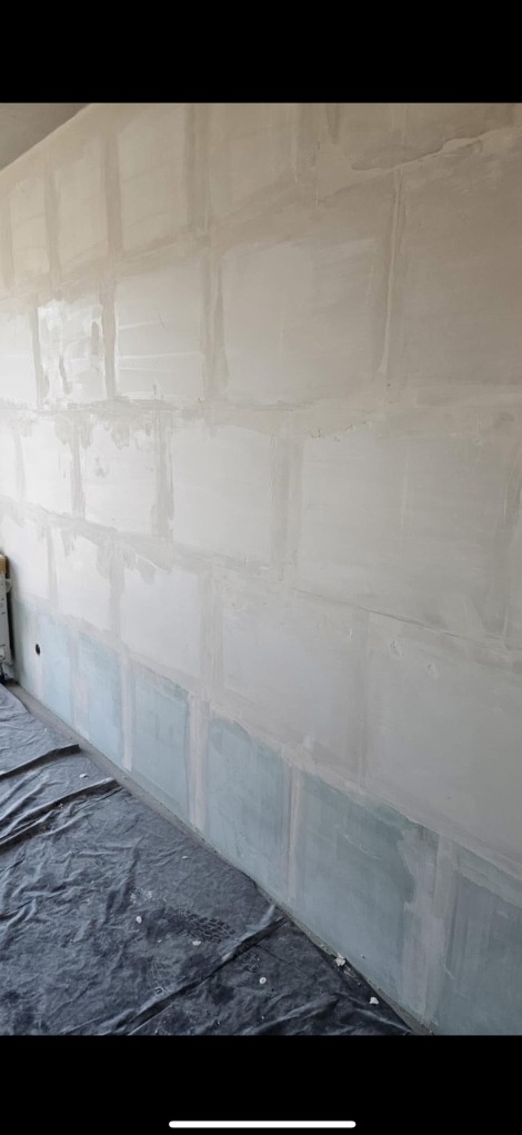 Ściana po szpachlowaniu, widoczne łączenia płyt kartonowo-gipsowych, przygotowana do dalszych prac wykończeniowych. Na podłodze folia ochronna.