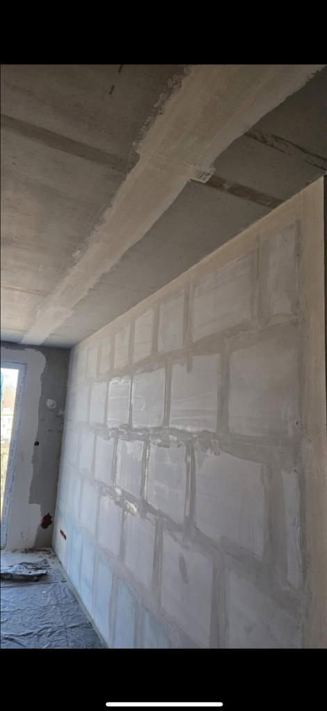 Ściana w trakcie szpachlowania z widocznymi kwadratowymi polami, surowy betonowy sufit, okno z lewej strony, folia ochronna na podłodze.