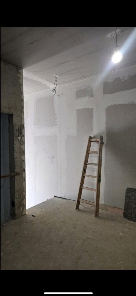 Pokój w trakcie szpachlowania ścian i sufitu, widoczna drewniana drabina oparta o ścianę. Surowy beton na podłodze i wystające kable z sufitu.