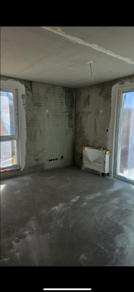 Pokój w trakcie prac wykończeniowych: surowe ściany, wylewka na podłodze, grzejnik zabezpieczony folią, okna z folią ochronną. Widoczne punkty elektryczne.