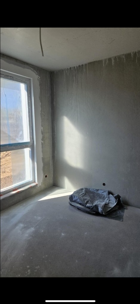 Pokój w stanie deweloperskim, ściany po szpachlowaniu, z widocznym oknem i kablem wystającym z sufitu. Na podłodze leży folia ochronna.