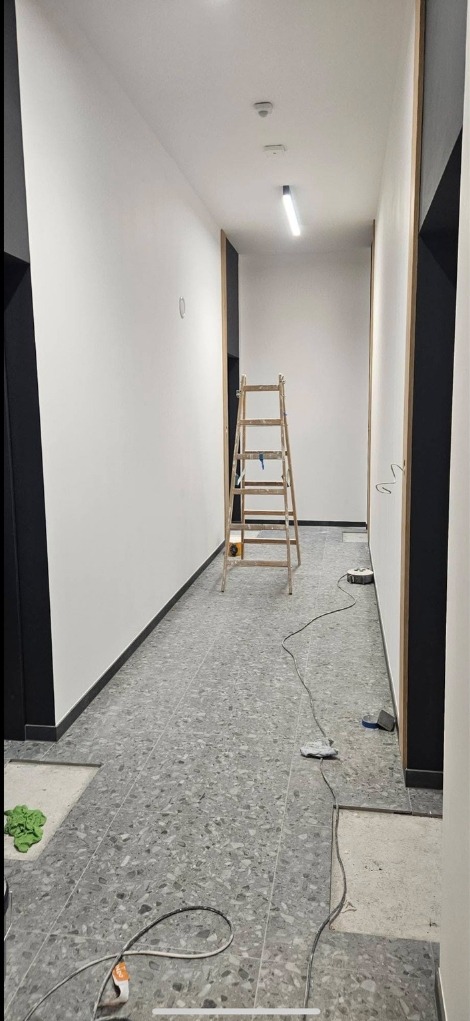 Korytarz w trakcie prac wykończeniowych: pomalowane ściany, położona podłoga z szarego lastryko, drabina oparta o ścianę. Widoczne kable i narzędzia.