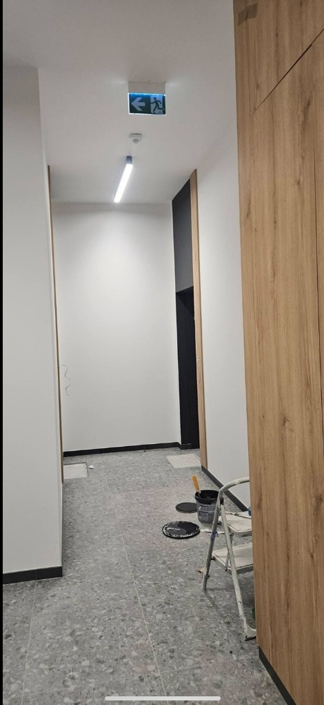 Korytarz w trakcie malowania: białe ściany, szara podłoga, drabina malarska, puszki z farbą. Widoczny znak ewakuacyjny i lampa sufitowa.