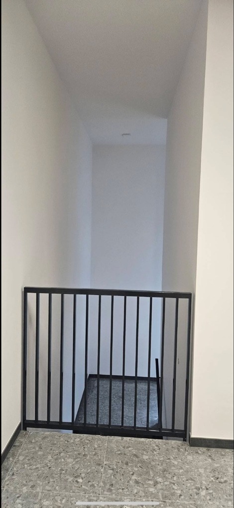 Nowoczesna, czarna barierka schodowa z metalowymi prętami na tle białych ścian i szarej podłogi z lastryko. Widok na schody w dół.