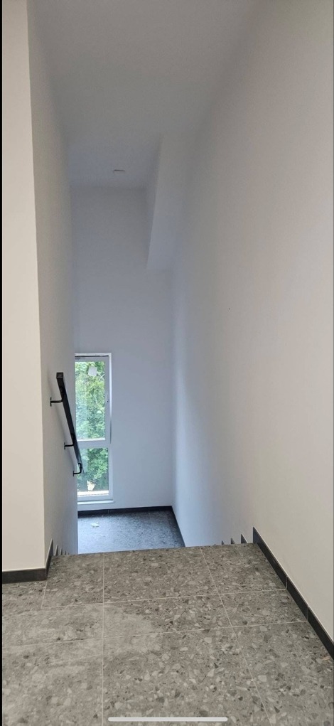 Wnętrze klatki schodowej po remoncie: szare płytki na podłodze, białe ściany, czarna poręcz przy oknie. Minimalistyczny design, świeżo po wykończeniu.