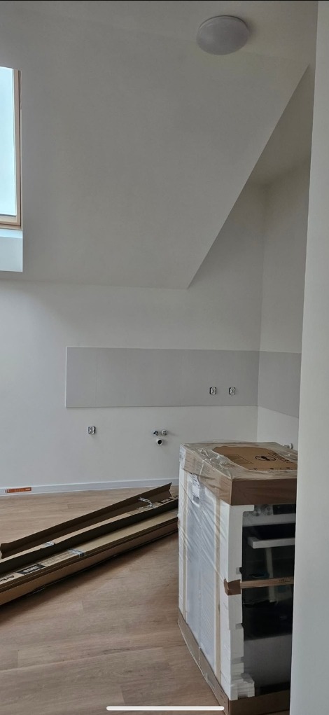 Wnętrze w trakcie wykańczania: podłoga, ściany przygotowane do malowania, częściowo rozpakowane meble kuchenne, okno dachowe. Etap przed ostatecznym wykończeniem.