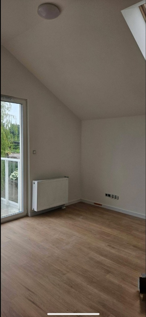 Jasne, skośne poddasze z drewnianą podłogą, białymi ścianami i grzejnikiem. Widok na zieleń przez balkonowe drzwi. Minimalistyczne wykończenie.