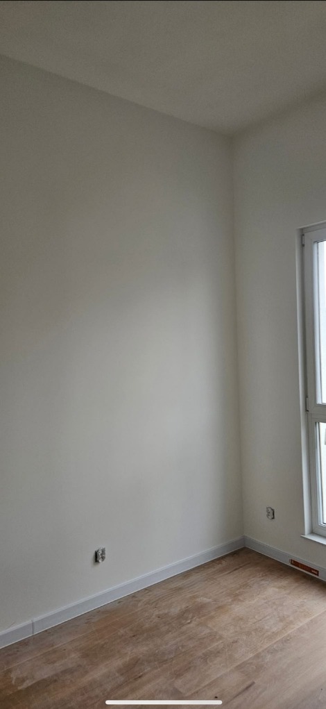 Jasne wnętrze po malowaniu: gładkie, kremowe ściany, drewnopodobna podłoga, biała listwa przypodłogowa i okno. Widoczne gniazdka elektryczne.