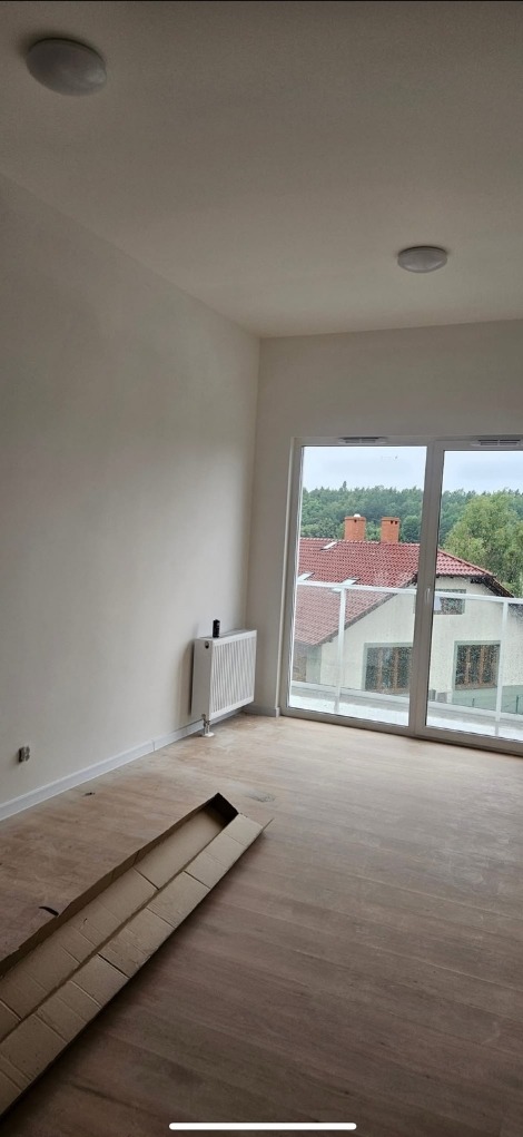 Jasne wnętrze po remoncie: świeżo pomalowane ściany, drewnopodobna podłoga, biały grzejnik i balkon z widokiem na domy z czerwonymi dachami.