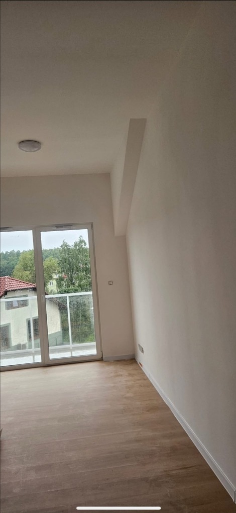 Jasne, odświeżone wnętrze z parkietem i dużym oknem balkonowym. Ściany pomalowane na biało, widoczny fragment sufitu i listwy przypodłogowe. Minimalistyczny styl.