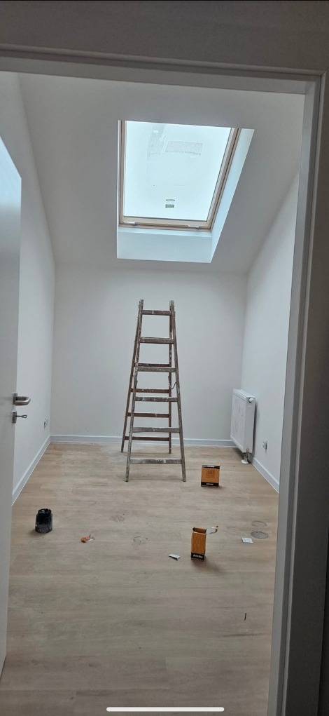Pokój po malowaniu z drewnianą drabiną, pudełkami po farbie i oknem dachowym. Podłoga wyłożona panelami. Białe ściany i listwy przypodłogowe.