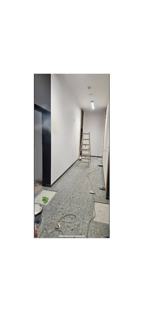 Wnętrze korytarza w trakcie odświeżania: pomalowane ściany na biało, szara podłoga z terazzo, drabina malarska, widoczne ślady prac remontowych.
