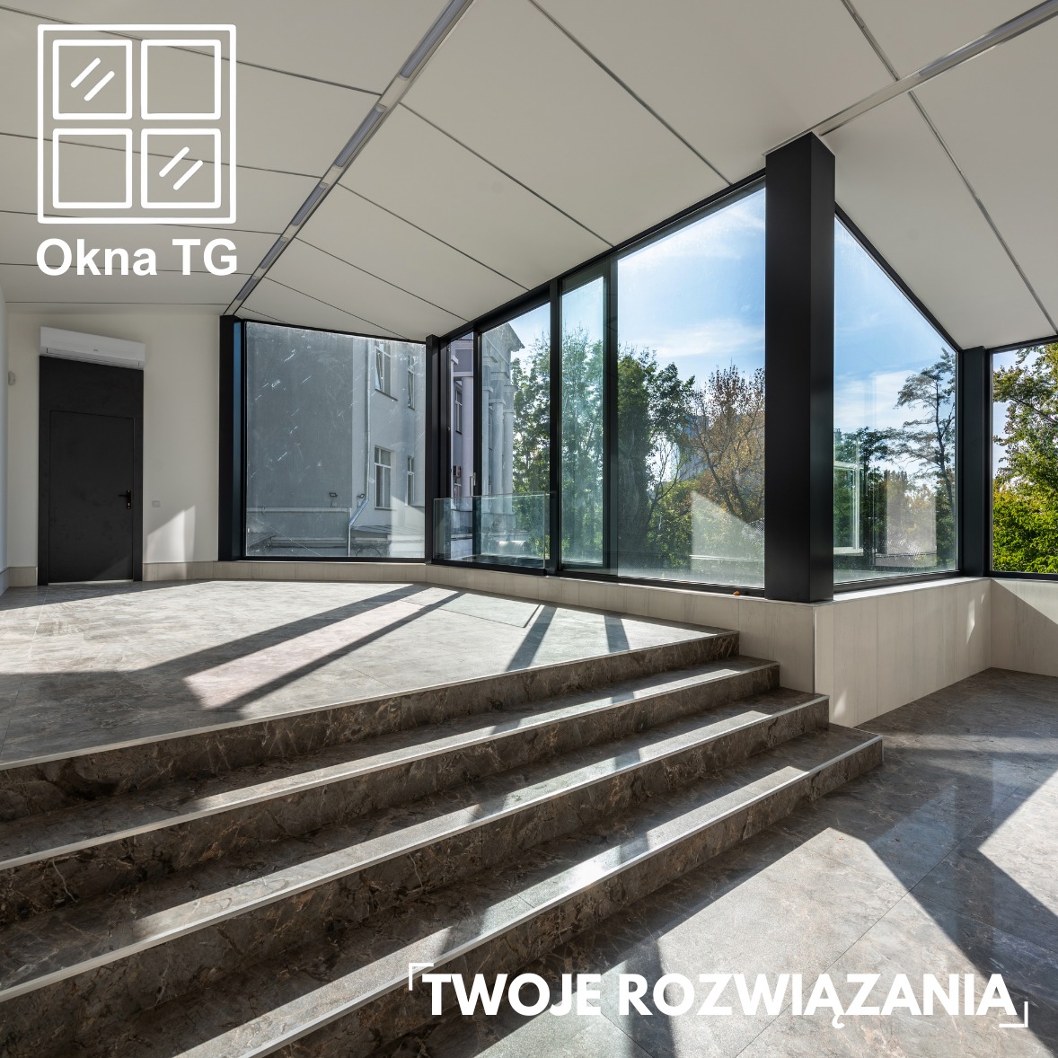 Przestronne wnętrze z dużymi oknami z czarnymi ramami, widok na zewnątrz, schody z płytek imitujących marmur. W tle czarne drzwi. Wnętrze doświetlone naturalnym światłem.