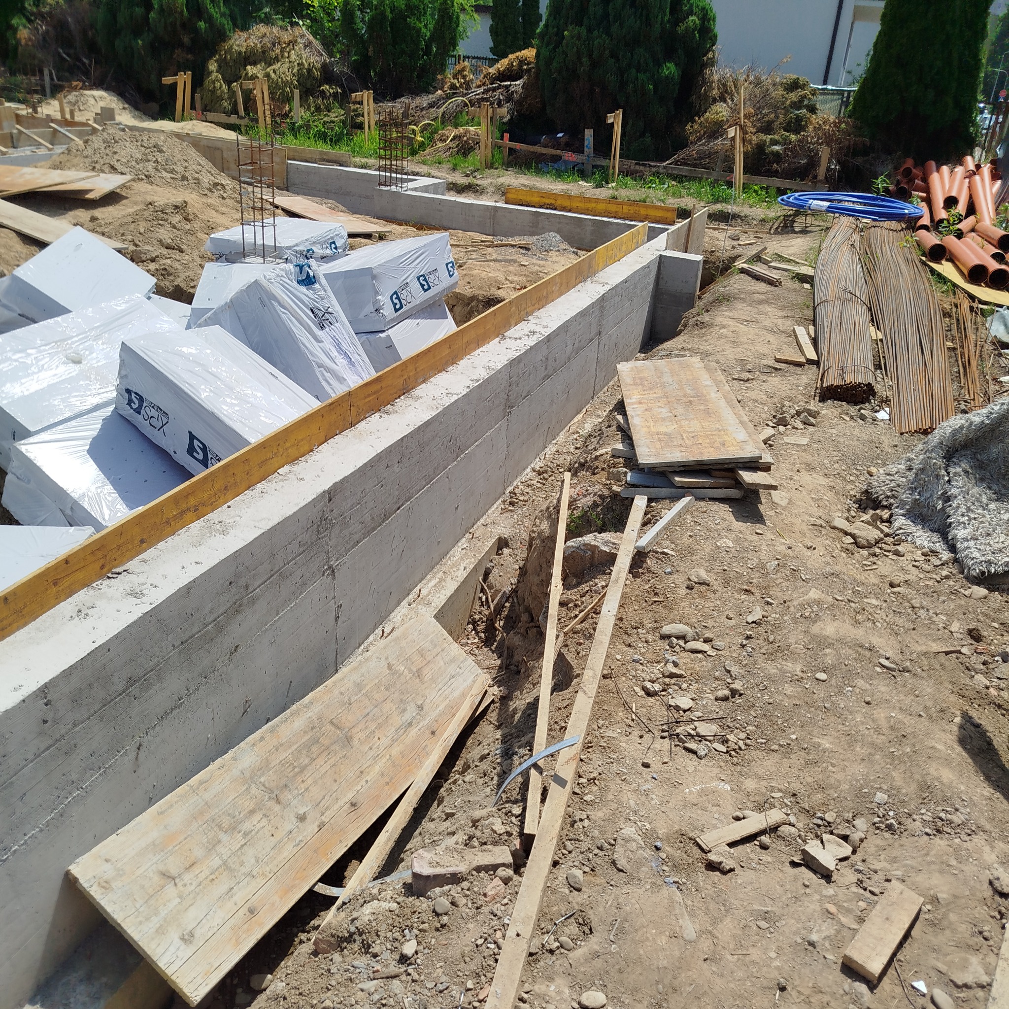 Wylane fundamenty z szalunkiem i zapakowanymi bloczkami styropianowymi, widoczne zbrojenia i rury. Prace ziemne w toku, widok z góry.