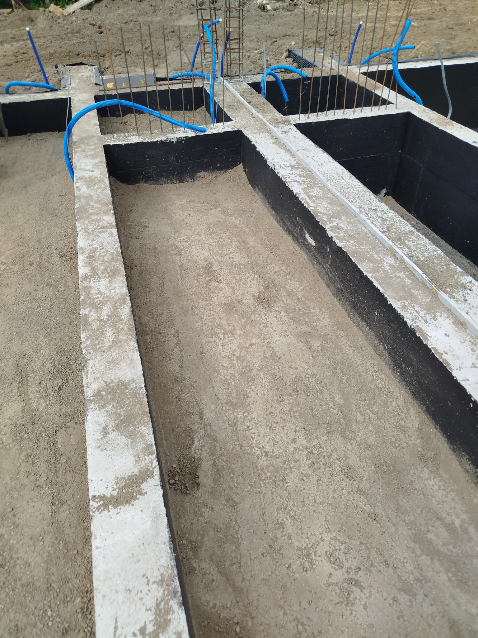 Zabezpieczone fundamenty z hydroizolacją i instalacją, widok z góry. Stalowe pręty zbrojeniowe wystają z betonowych ścian. Niebieskie rury instalacyjne w otworach.