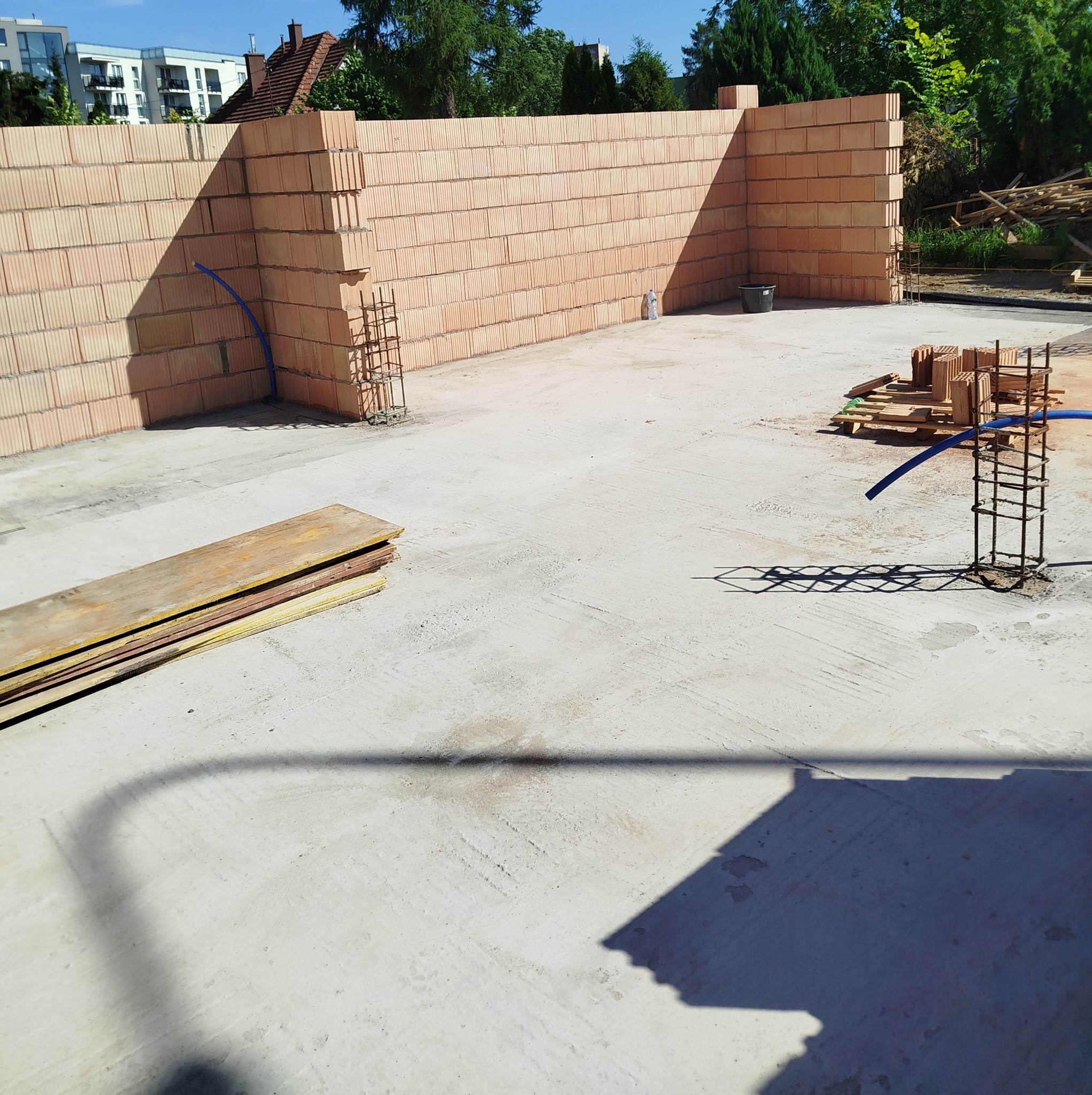 Wznoszone ściany z pustaków ceramicznych na fundamencie z widocznym zbrojeniem. Na podłożu leżą deski szalunkowe i pustaki. W tle budynki mieszkalne i zieleń.