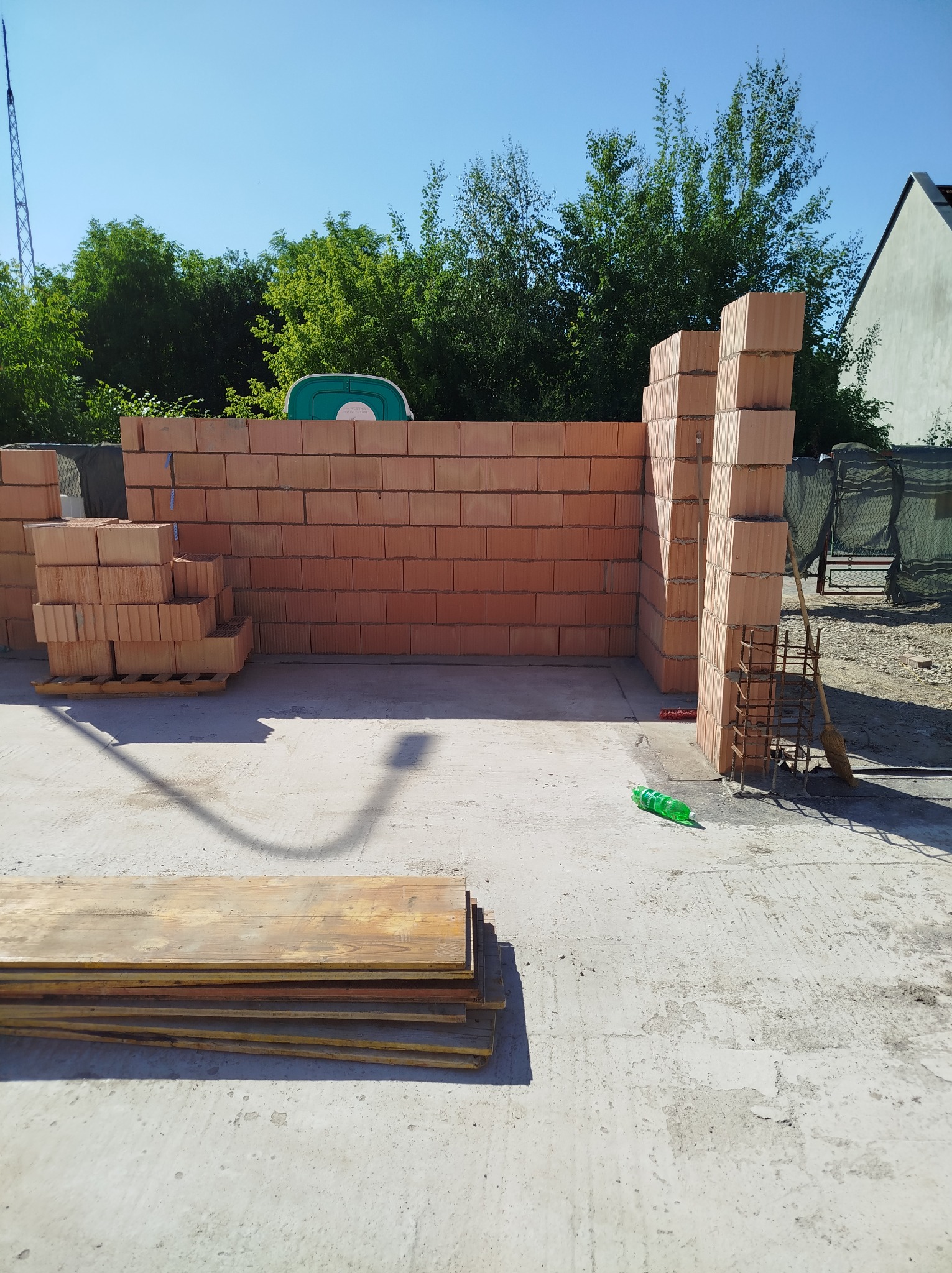 Wznoszenie muru z czerwonej cegły ceramicznej na fundamencie. Widoczne narzędzia murarskie, paleta z cegłami i przenośna toaleta w tle. Słoneczny dzień.