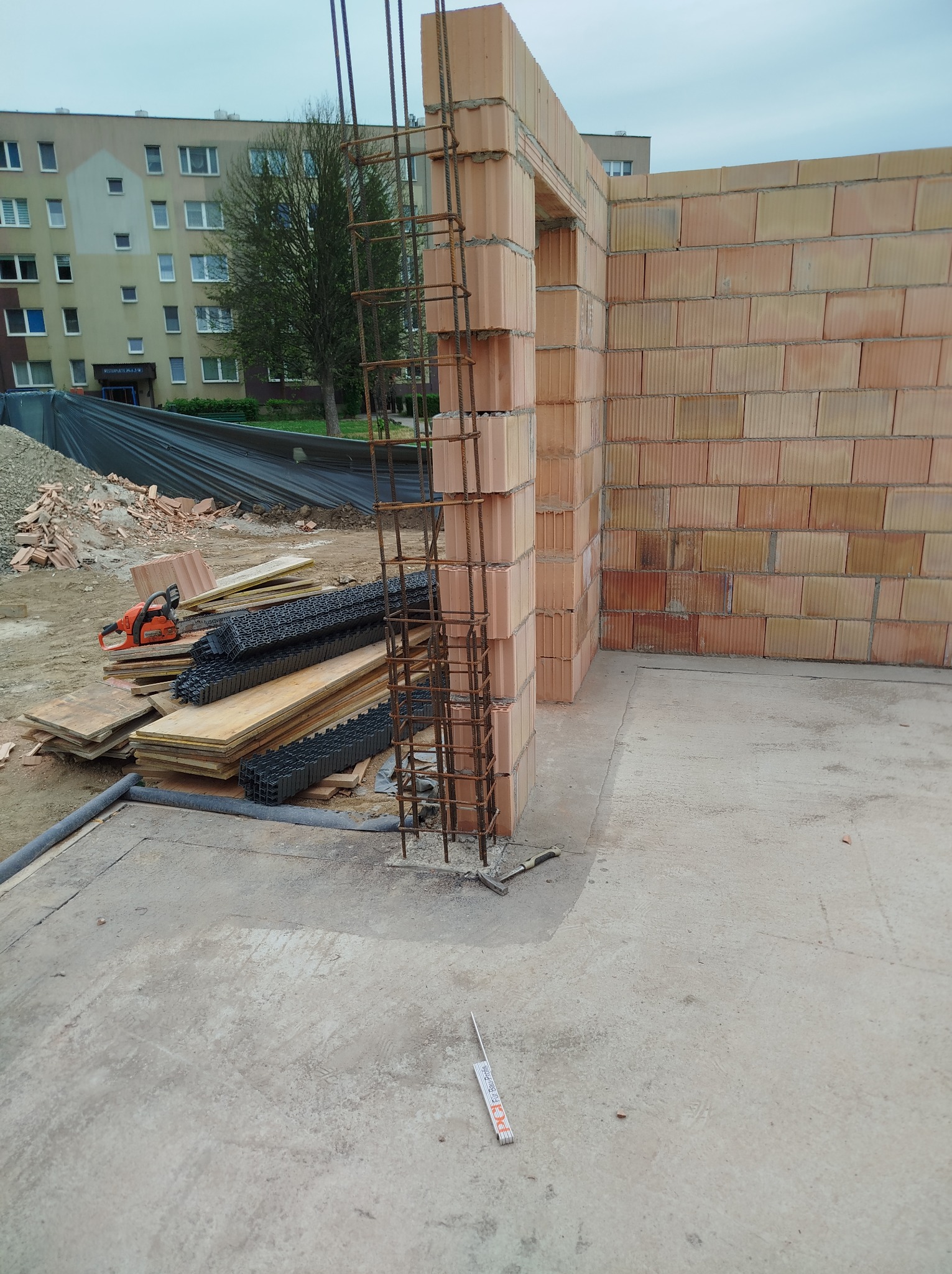 Wznoszenie ściany z pustaków ceramicznych Porotherm zbrojonej pionowo stalą. Widoczne narzędzia i materiały budowlane. Budynek mieszkalny w tle.