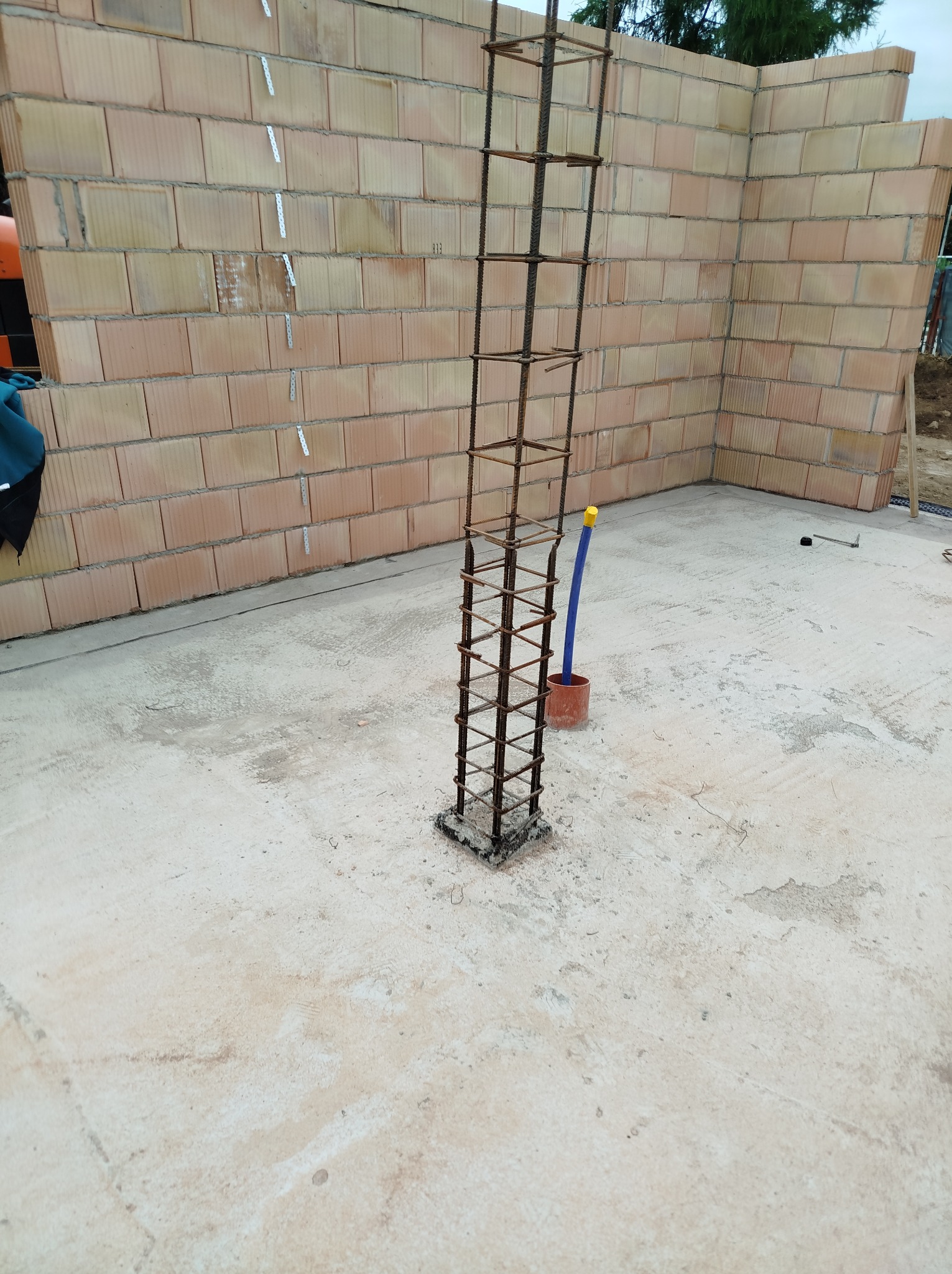 Zbrojenie pionowe na fundamencie, widoczne ściany z pustaków ceramicznych z metalowymi łącznikami. Etap budowy domu z niebieską rurką w pomarańczowej osłonie.