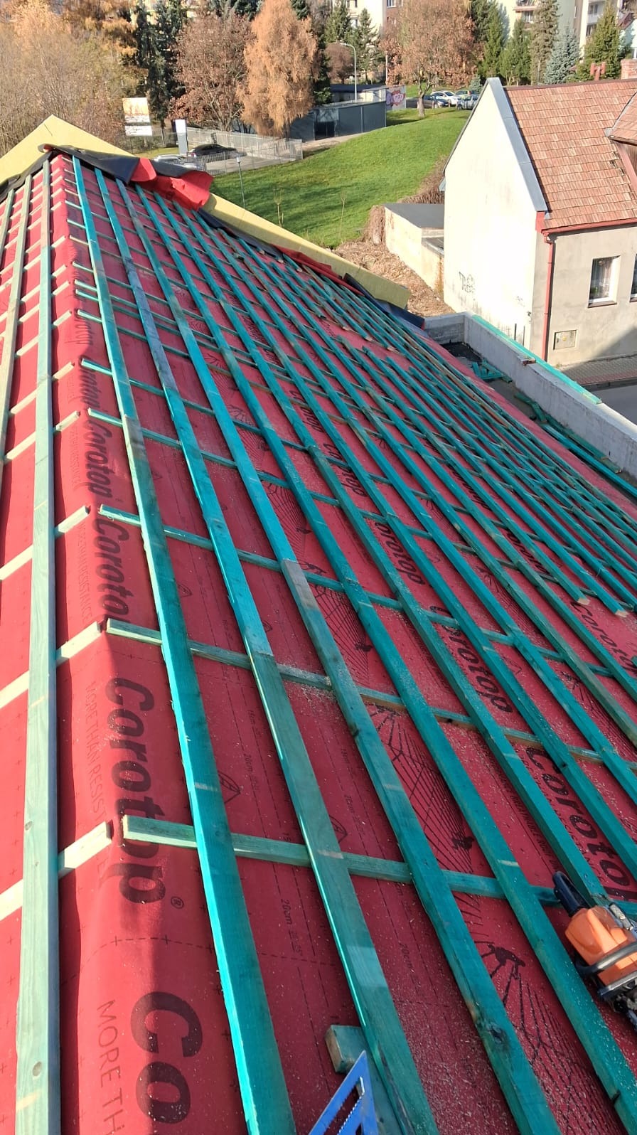 Czerwona membrana dachowa z zielonymi łatami, widok z góry na konstrukcję dachu w trakcie budowy, z narzędziami w prawym dolnym rogu i budynkami w tle.