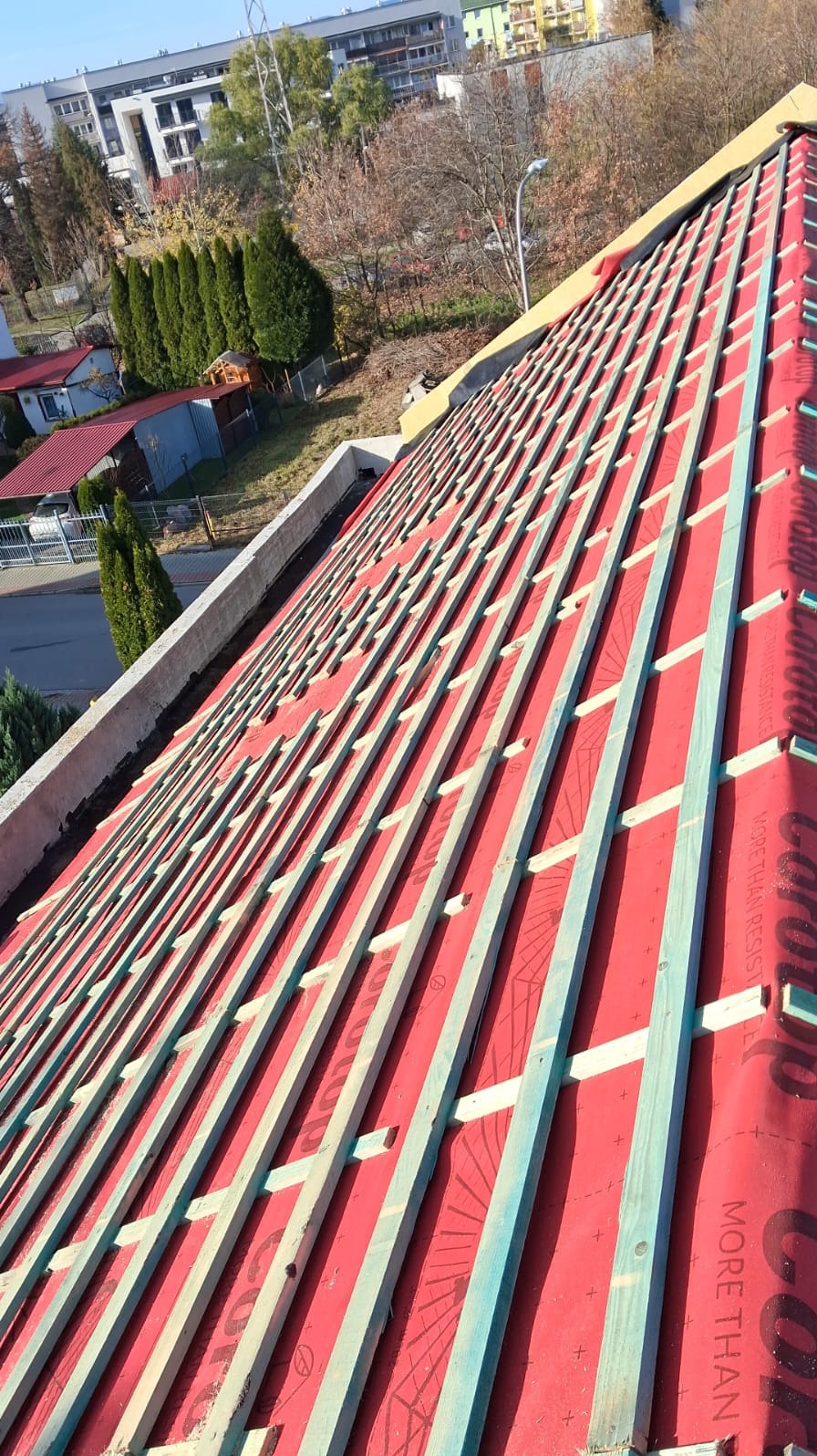 Czerwona membrana dachowa z drewnianymi łatami, widok z góry na budowę dachu. W tle zabudowa miejska i drzewa.