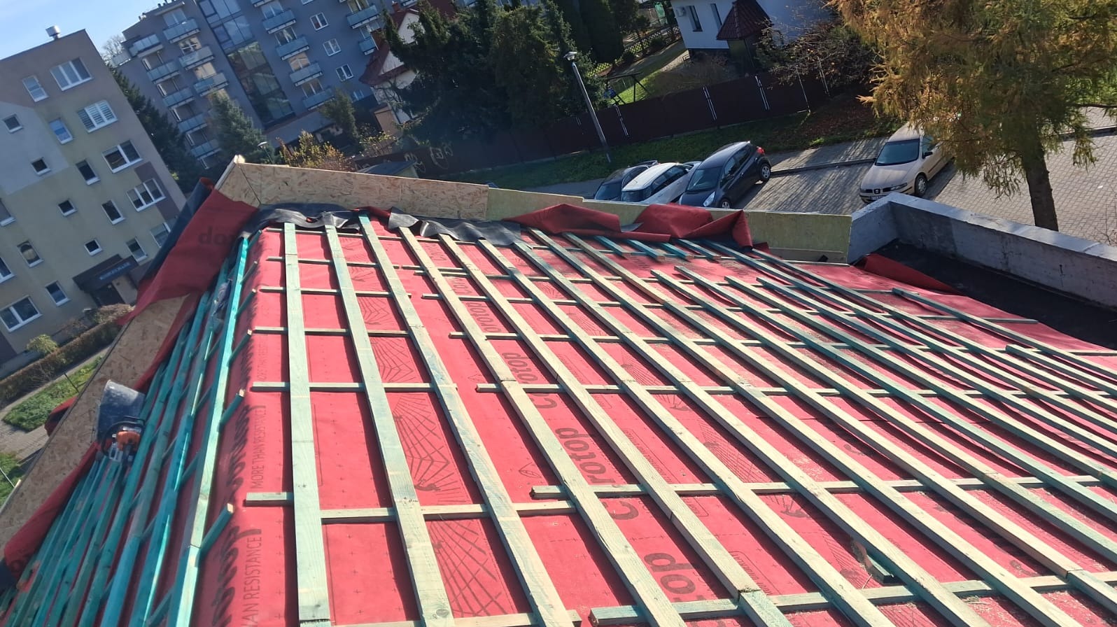 Czerwona membrana dachowa z drewnianą konstrukcją łat na tle miejskiego krajobrazu. Widoczne budynki mieszkalne i drzewa. Prace dekarskie w toku.