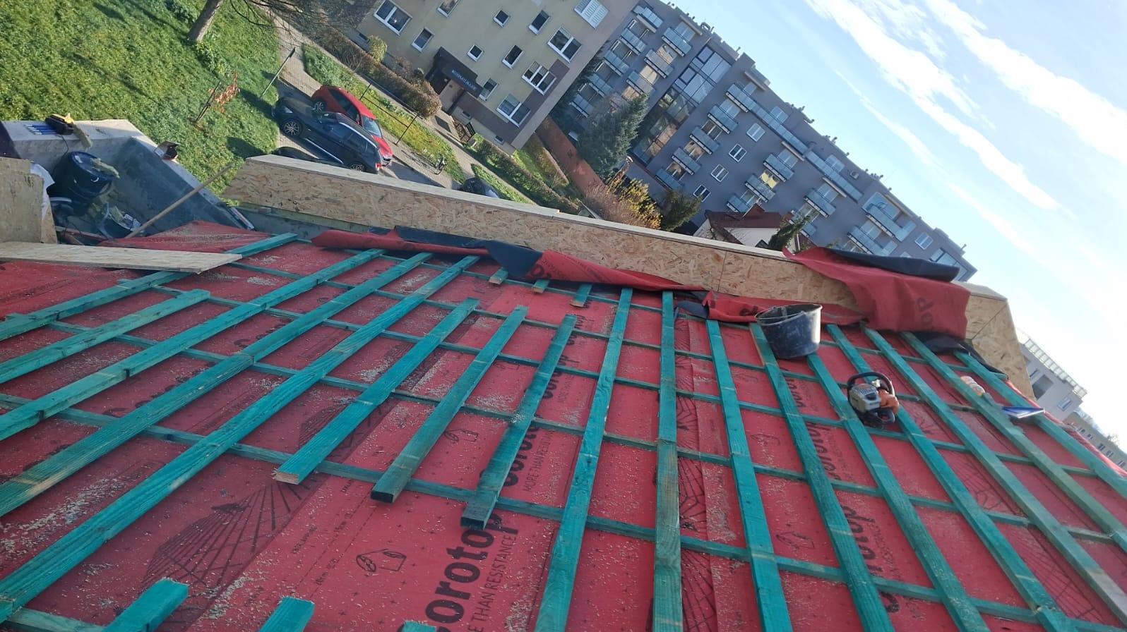 Konstrukcja dachu z łatami w kolorze turkusowym na czerwonej membranie dachowej, widok z góry na tle budynku mieszkalnego i nieba.