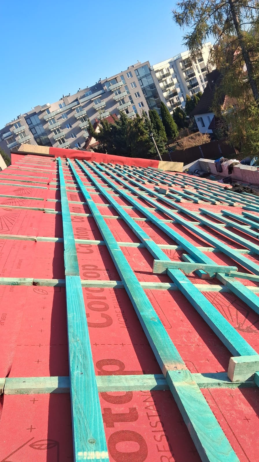 Czerwona membrana dachowa z niebieskimi łatami na tle budynku mieszkalnego i błękitnego nieba w słoneczny dzień. Widok z dołu na dach w trakcie budowy.