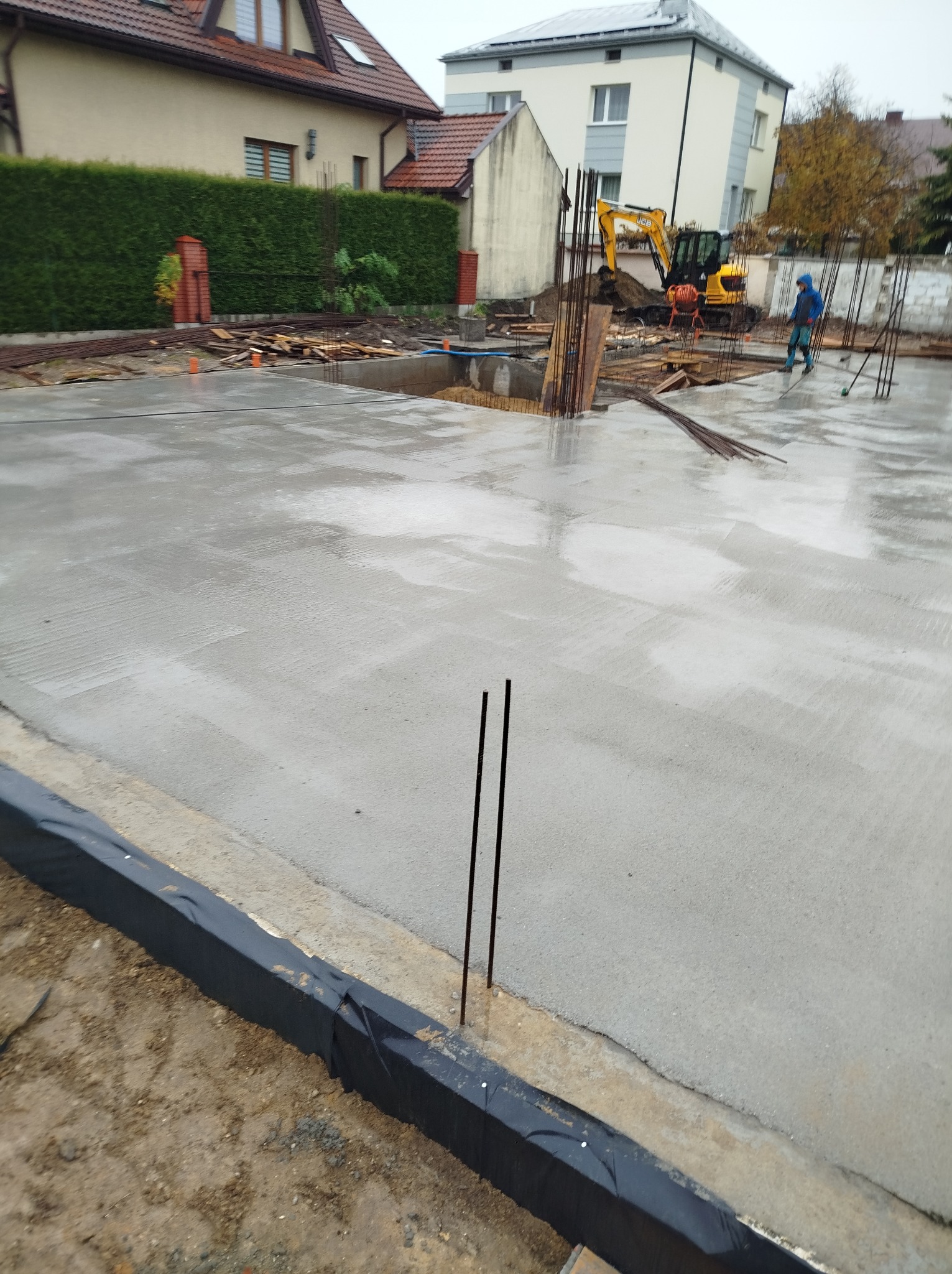 Zalana, świeża płyta fundamentowa z wystającymi prętami zbrojeniowymi. W tle koparka i pracownik budowlany. Widoczne zabezpieczenia hydroizolacyjne.