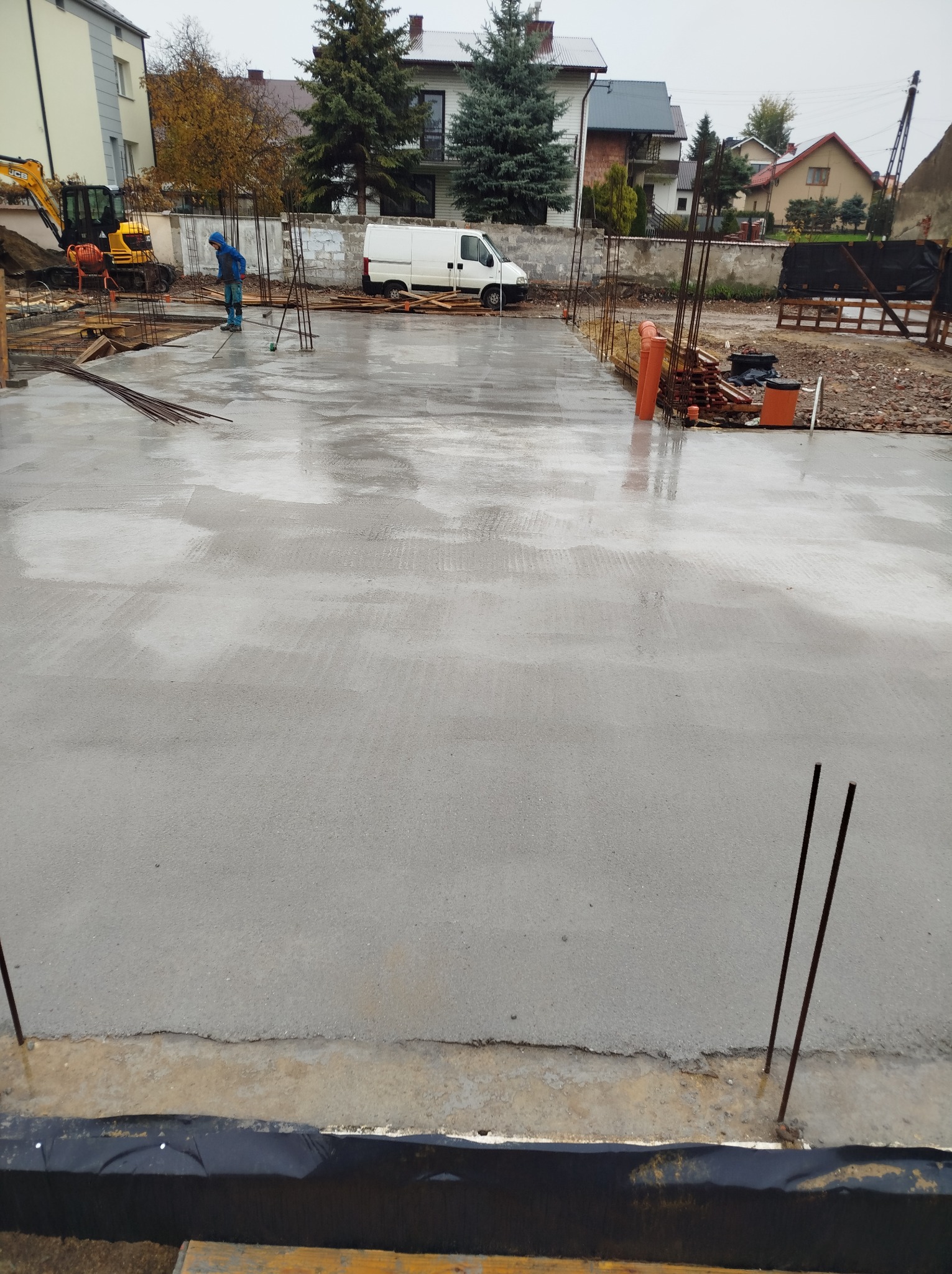 Wylana, świeża płyta fundamentowa z wystającymi prętami zbrojeniowymi. W tle budowa, koparka i biały bus. Praca w toku, widok z góry.