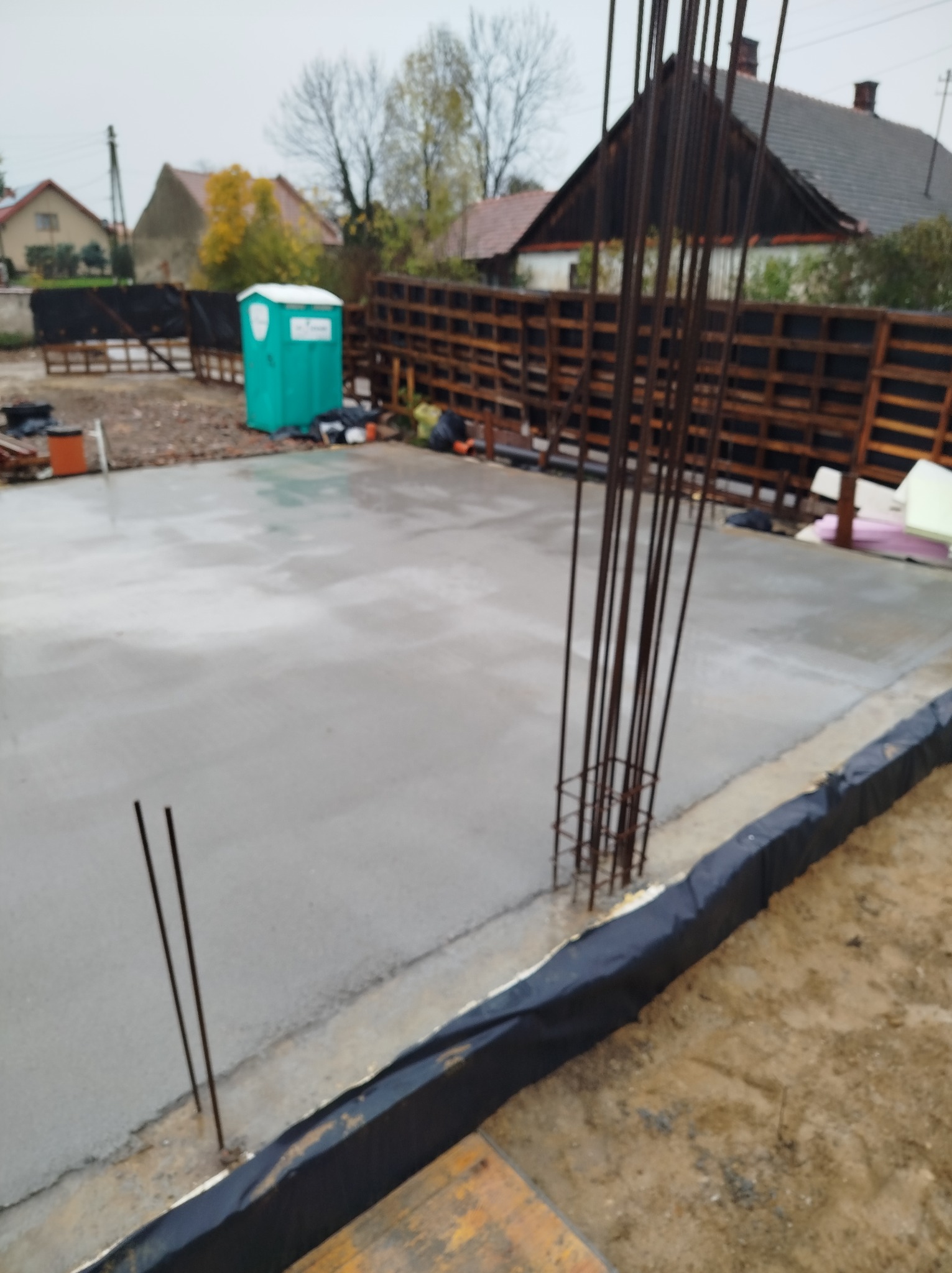Zalana, świeża płyta fundamentowa z wystającymi prętami zbrojeniowymi, zabezpieczona czarną folią. W tle ogrodzenie z palet i przenośna toaleta.