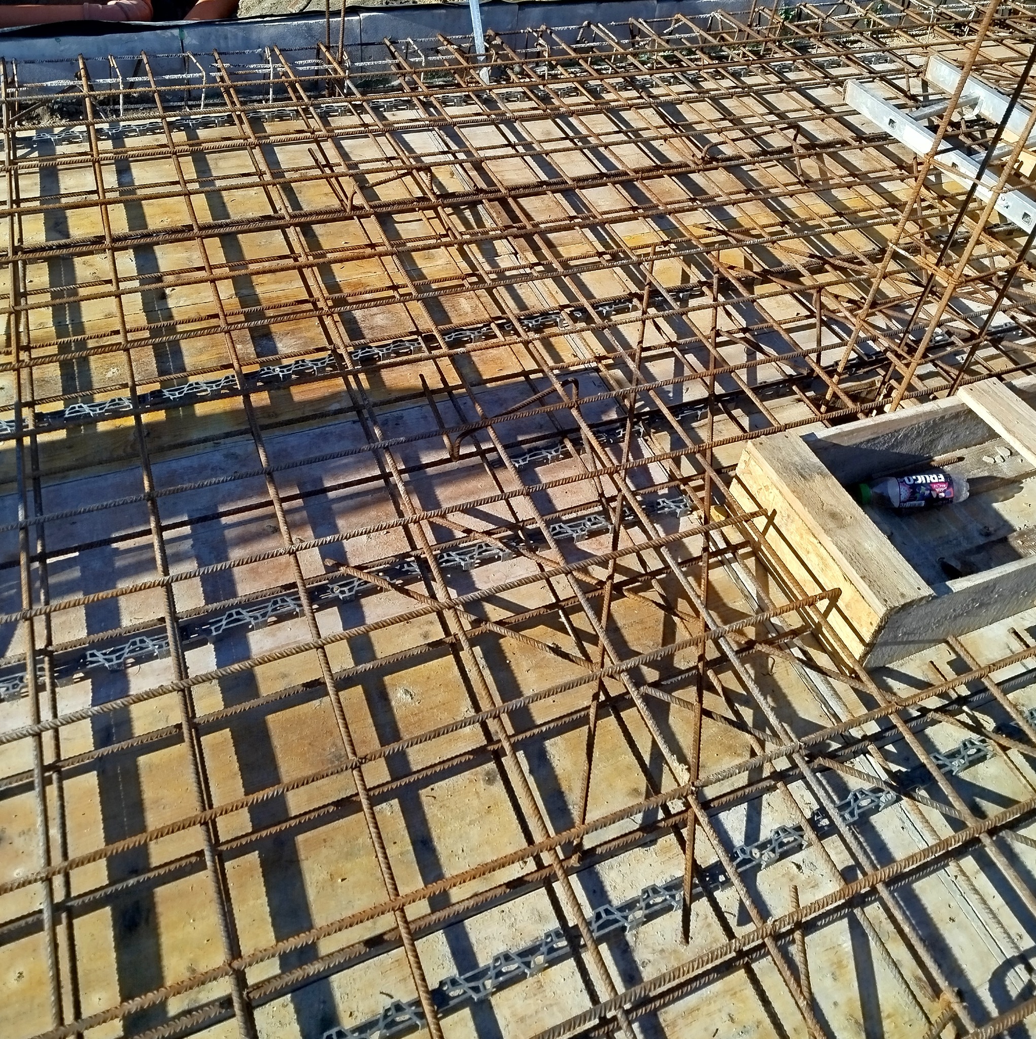 Zbrojenie płyty fundamentowej z widocznymi dystansami z tworzywa, przygotowane do zalania betonem. Widok z góry, ostre światło słoneczne.