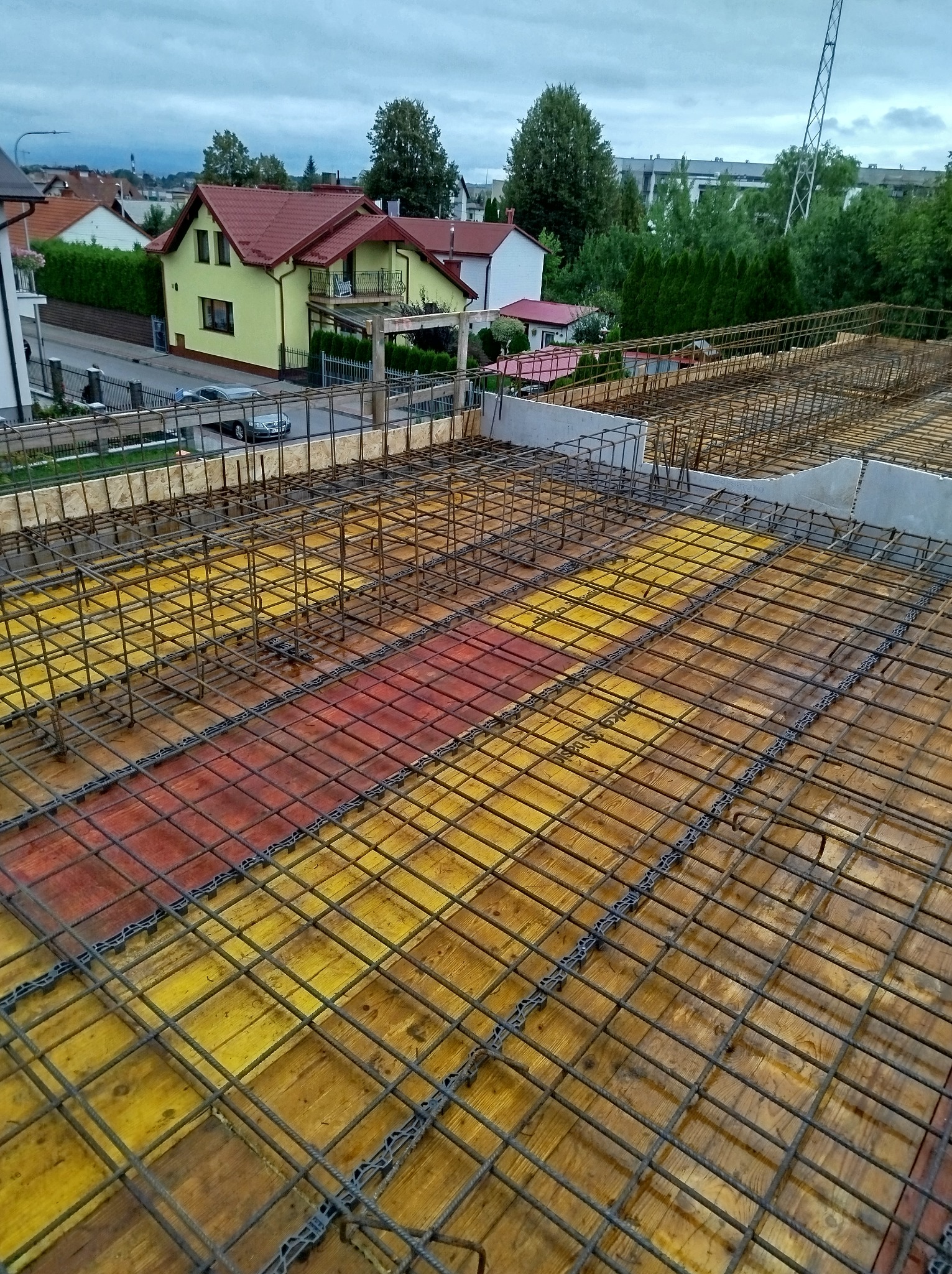 Zbrojenie stropu na deskowaniu z żółto-czerwonymi elementami, widok z góry. W tle domy jednorodzinne i drzewa pod pochmurnym niebem. Detal konstrukcji.